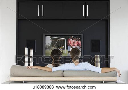 Stock Image - Back view of couple watching soccer game on television in living room. Fotosearch