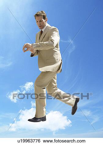 Stock Photograph - Business running whilst checking wristwatch in mid-air against cloudy sky. Fotosearch