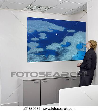 Stock Photograph - Contemplative young businesswoman looking at painting on wall in office. Fotosearch