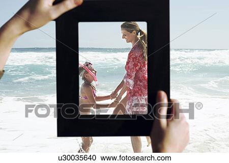 Stock Image - Female hands framing mother and daughter playing in water at beach. Fotosearch