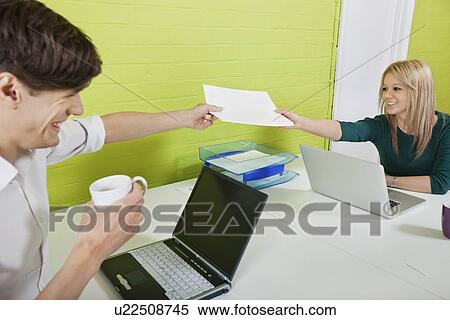 Stock Photography - Happy young businesspeople passing documents to each other with laptops at desk. Fotosearch