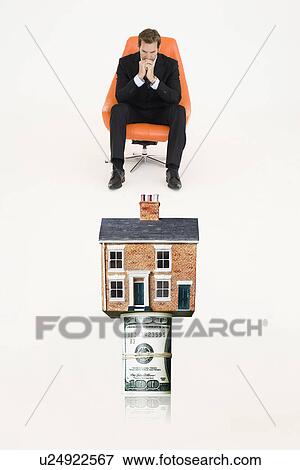 Stock Photo - House on top of roll of bills with pensive businessman on chair representing expensive real estate. Fotosearch