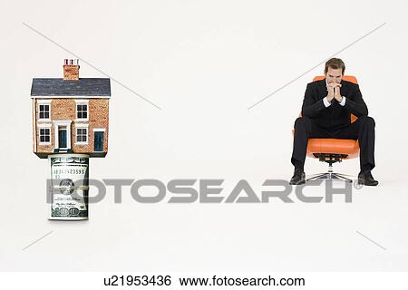 Stock Photograph - House on top of roll of bills with pensive businessman on chair representing expensive real estate. Fotosearch