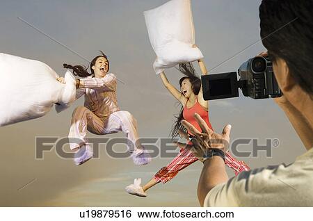 Stock Photograph - Man video recording two playful women in sleepwear having a pillow fight. Fotosearch
