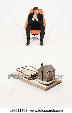 Stock Photo - Model home on mouse trap with worried businessman sitting on chair representing increasing real estate rates. Fotosearch
