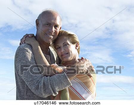 Stock Photograph - Portrait of smiling senior couple hugging each other against cloudy sky. Fotosearch