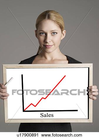 Stock Image - Portrait of young businesswoman showing sales graph against gray background. Fotosearch