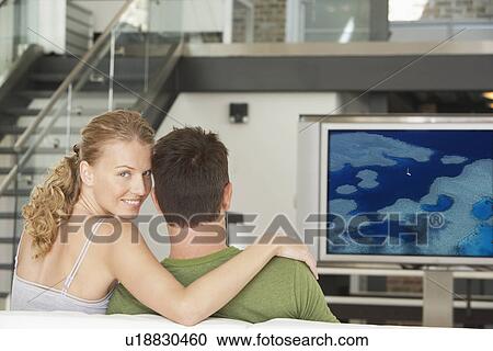 Stock Image - Portrait of young Caucasian woman with man watching movie on television in living room. Fotosearch
