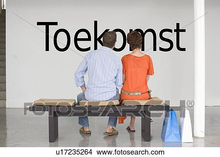 Picture - Rear view of couple seated on bench reading Dutch text "Toekomst" (future) on wall. Fotosearch