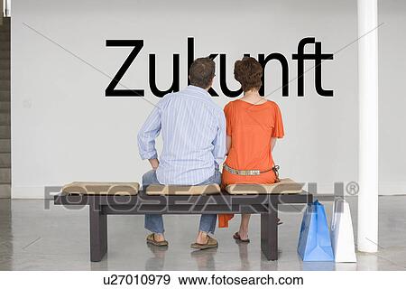 Stock Photo - Rear view of couple seated on bench reading German text "Zukunft" (future) on wall. Fotosearch