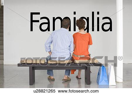 Stock Photography - Rear view of couple seated on bench reading Italian text "Famiglia" (family) on wall. Fotosearch