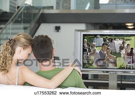 Stock Image - Rear view of young couple watching television in living room. Fotosearch