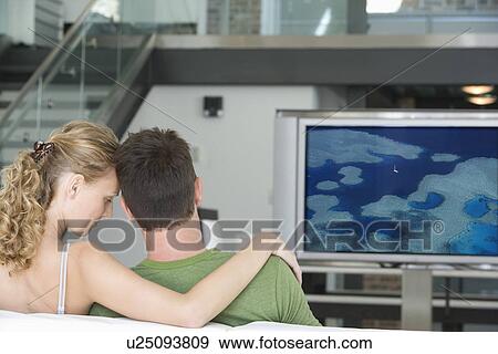Rear view of young couple watching television in living room View Large Photo Image Stock Photo - Rear view of young couple watching television in living room. Fotosearch
