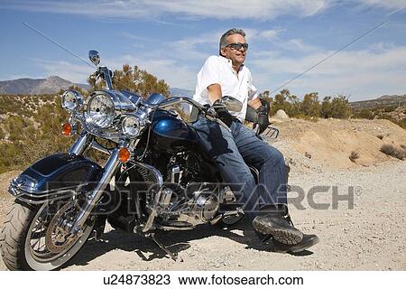 Stock Image - Senior man leaning on motorcycle on desert road. Fotosearch