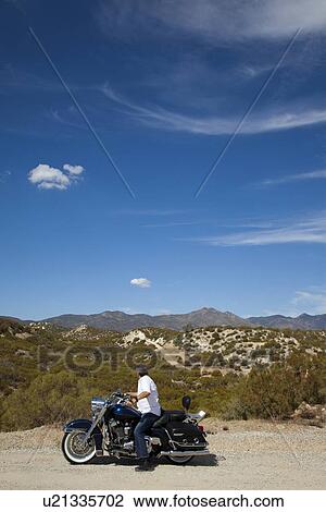 Stock Image - Senior man riding motorcycle on desert road looking to mountains in background. Fotosearch