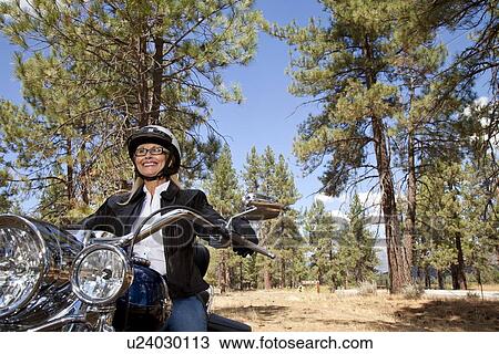 Stock Image - Senior woman riding motorcycle through a forest. Fotosearch