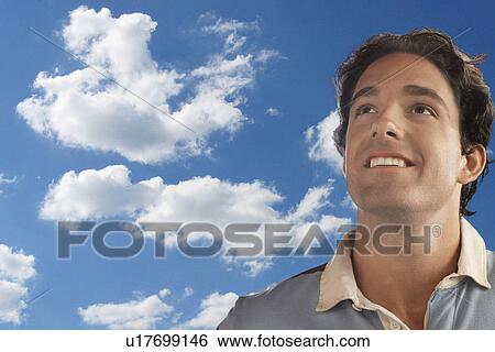 Stock Photograph - Smiling young man daydreaming against cloudy sky. Fotosearch