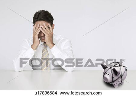 Picture - Worried businessman at table with trapped piggy bank representing financial difficulties. Fotosearch
