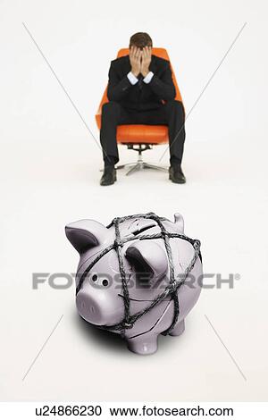 Stock Image - Worried businessman on chair and piggybank tied with rope representing financial difficulties. Fotosearch