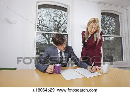 Stock Photo - Young businesswoman looking at male colleague signing documents at conference table. Fotosearch