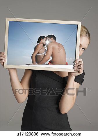 Stock Photograph - Young businesswoman peeking from behind photograph of couple at beach against gray background. Fotosearch