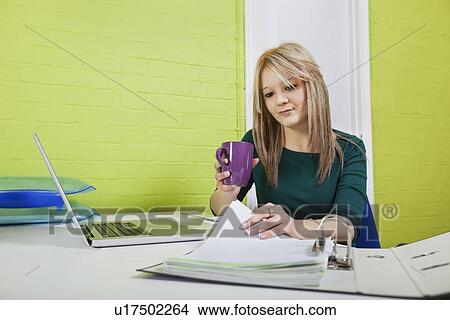 Picture - Young businesswoman with coffee mug reviewing documents at desk. Fotosearch