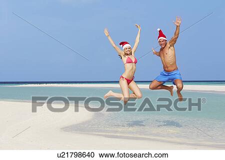 Stock Image - Couple Jumping On Beach Wearing Santa Hats. Fotosearch