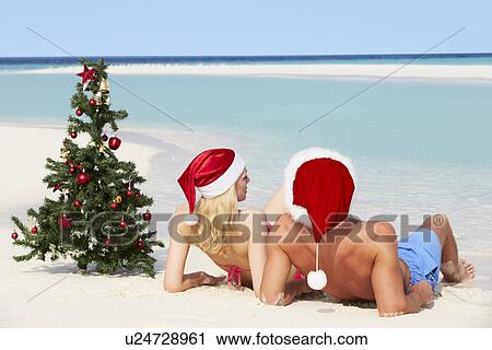 Stock Image - Couple Sitting On Beach With Christmas Tree And Hats. Fotosearch