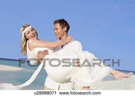 Stock Image - Groom Carrying Bride At Beautiful Beach Wedding. Fotosearch