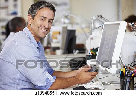 Stock Image - Man Using Mobile Phone At Desk In Busy Creative Office. Fotosearch