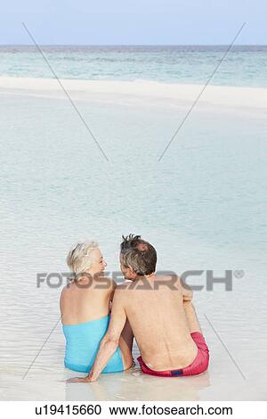 Stock Image - Rear View Of Senior Couple Sitting On Beautiful Beach. Fotosearch