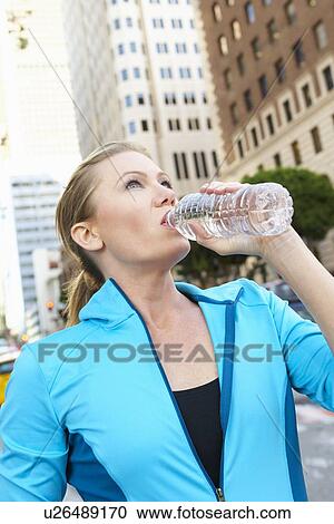 Stock Image - Woman Drinking Water Whilst Running Along Urban Street. Fotosearch