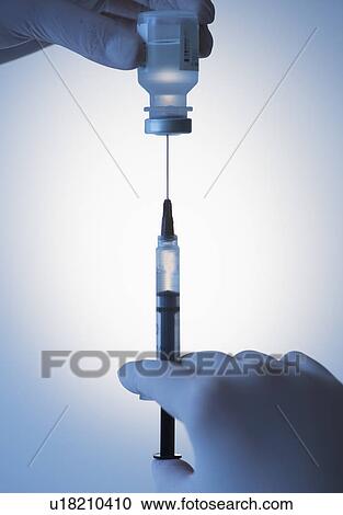 Stock Image - Close up of hands in surgical gloves preparing syringe for injection, studio shot. Fotosearch