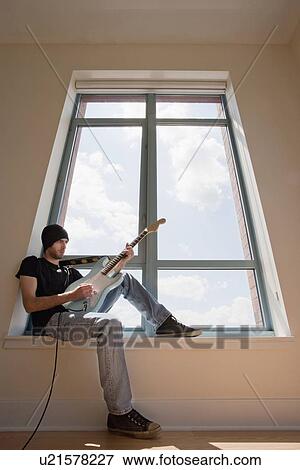 Stock Photo - Low angle view of young man sitting on window sill and playing electric guitar. Fotosearch