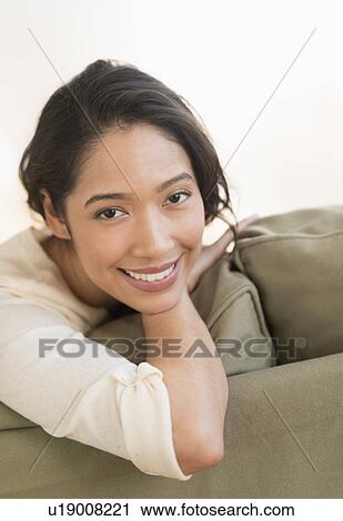Stock Image - Portrait of young woman sitting on couch. Fotosearch