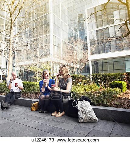 Stock Image - Businesswomen sitting on wall for coffee break. Fotosearch