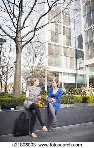 Stock Image - Businesswomen sitting on wall for coffee break. Fotosearch