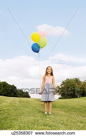 Girl holding balloons standing on grass View Large Photo Image Stock Photo - Girl holding balloons standing on grass. Fotosearch