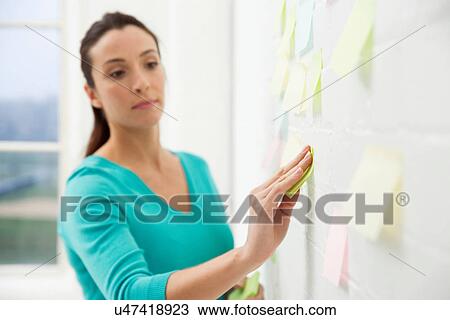 Stock Image - Mid adult woman sticking adhesive notes to wall. Fotosearch