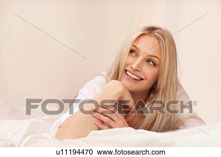 Stock Image - Portrait of a young blond woman lying on bed smiling. Fotosearch