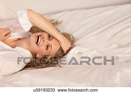 Stock Image - Portrait of a young blond woman lying on bed laughing. Fotosearch
