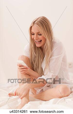 Stock Image - Portrait of a young blond woman sitting on bed using smartphone. Fotosearch
