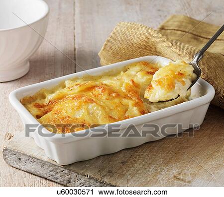 Stock Image - Potato gratin in white serving dish with metal spoon. Fotosearch