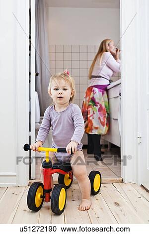 Stock Image - Toddler girl playing on tricycle, mother in background. Fotosearch