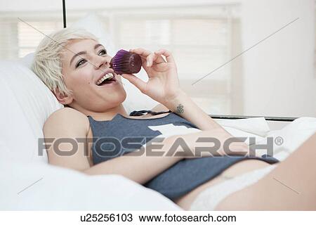 Stock Image - Woman lying on bed eating cupcake. Fotosearch