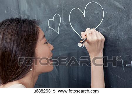 Stock Photography - Young woman drawing heart on blackboard. Fotosearch
