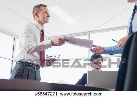 Stock Image - Businessman handing colleague document in office. Fotosearch
