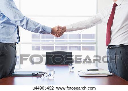 Stock Image - Businessmen shaking hands above desk. Fotosearch