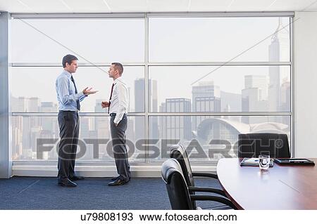 Businessmen standing by office window talking View Large Photo Image Stock Image - Businessmen standing by office window talking. Fotosearch