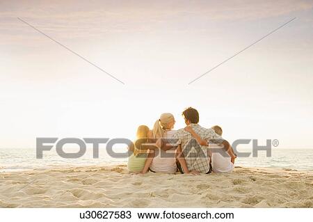 Stock Image - Family sitting on beach with arms around each other. Fotosearch